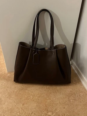 Den virala mörkbruna väskan från zara  - Stilren och rymlig mörkbrun shoppingväska i slätt skinn med vita kontrastsömmar och dubbla axelband. Väskan har en enkel tag-detalj framtill och är perfekt för dig som vill ha plats för allt du behöver under dagen. Klassisk design som funkar till många looks.