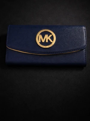 Mörkblå plånbok från Michael Kors - Snygg mörkblå plånbok från Michael Kors med guldfärgad logga på framsidan. Plånboken har flera fack, dragkedja inuti och stängs med tryckknapp. Utsidan är i skinn och insidan är beige med detaljer i brunt och MK-mönster.