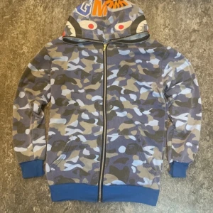 BAPE blå camo hoodie med shark-hood - Säljer en ikonisk BAPE hoodie i blått camo-mönster med hel dragkedja och shark-motiv på huvan. Tröjan har blå muddar och fickor framtill. Färgglada bokstäver och tigertryck på huvan ger en unik streetwear-look.
