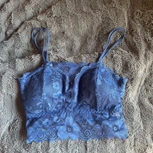 Blå spetsig linnetopp med blommönster - Supersöt blå linnetopp i spets med blommigt mönster och tunna axelband. Toppen är croppad och har fina vågiga kanter nertill. Perfekt för dig som gillar romantisk och feminin stil.