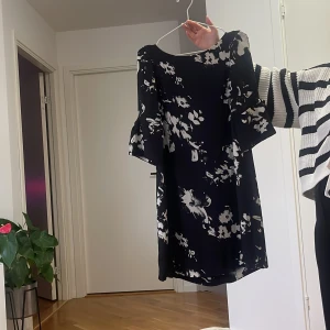 Svart blommig kortklänning - Svart kortklänning med vita blommiga mönster och vida, korta ärmar. Klänningen har en avslappnad passform och är perfekt för dig som gillar en stilren men ändå lekfull look. Materialet känns lätt och flowy.