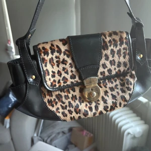 Leopardmönstrad axelväska i skinn - Cool axelväska med leopardmönstrad framsida och detaljer i svart skinn. Väskan har ett snyggt guldigt lås framtill och bred axelrem. Perfekt storlek för det viktigaste och ger en edgy vibe till din outfit.