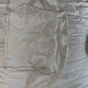 Vit stickad tröja med mönster - Mysig vit stickad tröja med grovt kabelstickat mönster framtill och på ärmarna. Tröjan har rund halsringning och ribbade muddar vid ärmslut och nederkant. Perfekt för kyliga dagar och enkel att matcha med jeans eller kjol.