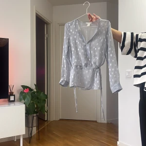 Ljusblå omlottblus med knyt - Säljer en ljusblå blus med vita prickar och omlottmodell. Blusen har v-ringning, långa ärmar och knyt i midjan för en snygg passform. Tunt och lätt material som ger en luftig känsla.