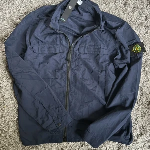 Marinblå vindjacka från Stone Island - Säljer en marinblå vindjacka från Stone Island med dragkedja framtill och två bröstfickor. Jackan har klassisk logotyp-patch på ärmen och är tillverkad i lätt polyester. Stor i storleken och pris kan diskuteras