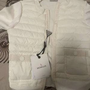 Vit quiltad dunväst från Moncler - Snygg vit dunväst från Moncler med quiltad framsida, två fickor med knappdetalj och dragkedja. Västen har en stilren look och är tillverkad i polyester och dun, perfekt för lager-på-lager. Klassisk design med Moncler-logga på etiketten.