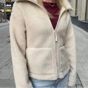 Beige teddyjacka med dragkedja - Mysig beige teddyjacka med dragkedja framtill och stora fickor. Jackan har en hög krage och är perfekt för kyliga dagar. Den är långärmad och har en avslappnad passform som passar till det mesta.