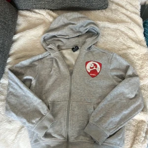 Grå Nike hoodie med Nobel-tryck - Grå hoodie från Nike med dragkedja och huva. På bröstet finns ett rött Nobel-tryck. Tröjan har två fickor framtill och är tillverkad i mjuk bomullsblandning. Perfekt för chill dagar eller när du vill ha en sportig look.