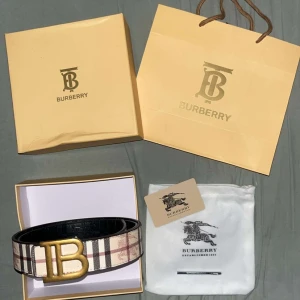 Burberry bälte med guldspänne  - Säljer ett Burberry bälte med klassiskt rutigt mönster i beige, svart, vitt och rött. Bältet har ett stort, guldigt 'B'-spänne och är gjort i skinn. Perfekt accessoar för att lyfta din outfit med en lyxig touch. Bälte har 2 sidor som man kan använda 