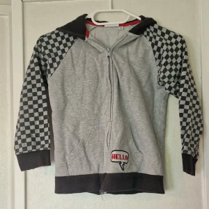 Grå hoodie med rutiga ärmar KappAhl - Grå hoodie från KappAhl med svarta och grå rutiga ärmar, svart huva och muddar. Dragkedja framtill och broderad detalj med texten 'HELLO' på magen. De skavanker som jag har kunnat se är några trådar som letat sig lösa och tygmärket som är nopprigt (se bild).