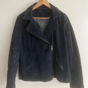 Mörkblå mockajacka med dragkedjor - Snygg mörkblå mockajacka med klassisk bikerkrage och sned dragkedja framtill. Jackan har två fickor med dragkedjor och långa ärmar med dragkedjedetaljer vid ärmsluten. Perfekt för dig som vill ha en stilren och cool look. Nästan inte använd