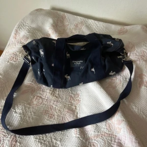 Mörkblå axelväska Abercrombie & Fitch - Snygg mörkblå axelväska från Abercrombie & Fitch med vita älgmönster. Väskan har både handtag och justerbar axelrem, samt dragkedja upptill. Tillverkad i slitstark bomull, perfekt för att bära med sig allt man behöver. Märkeslogga framtill.