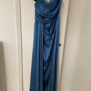 Blå långklänning med drapering - Elegant blå långklänning i glansigt material med v-ringning och draperad framsida. Klänningen har offshoulder-ärmar och en figurnära passform upptill som faller snyggt längs kroppen. Perfekt för bal eller festliga tillfällen.