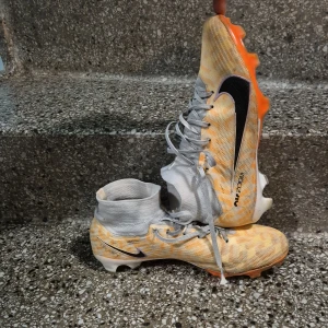 Nike Mercurial Zoom Superfly 9 Elite NU FG 'Guava Ice' DZ3457-800 - Säljer ett par Nike Air Zoom fotbollsskor med högt skaft och snörning. Skorna har ett mönstrat mesh-överdrag i orange och vitt, svart Nike-logga och orange sula med dobbar. Perfekta för dig som vill sticka ut på planen.