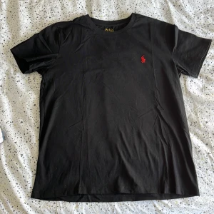 Svart t-shirt Polo Ralph Lauren S - Svart t-shirt från Polo Ralph Lauren i slim fit-modell. Klassisk design med korta ärmar och rund hals. T-shirten har det ikoniska röda broderade Polo-loggan på bröstet. Tillverkad i mjuk bomull för en skön känsla.
