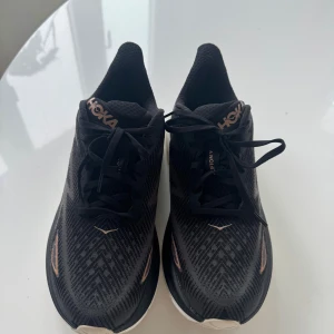 Svarta Hoka Clifton 9 sneakers - Säljer ett par svarta Hoka Clifton 9 sneakers med rosa detaljer och vit sula. Skorna har snörning och är tillverkade i ett ventilerande syntetmaterial med mönstrad ovandel. Perfekta för dig som gillar att springa eller promenera långt.