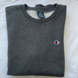 Mörkgrå sweatshirt från Champion - Mörkgrå sweatshirt från Champion med klassisk rund hals och broderad logga på bröstet. Tröjan har långa ärmar och är tillverkad i mjuk bomullsblandning, perfekt för chill dagar eller när du vill ha en clean och enkel stil.