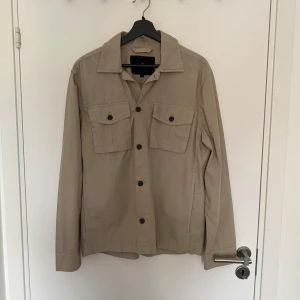 Beige overshirt från Morris Stockholm - Stilren beige overshirt från Morris Stockholm med klassisk krage, två bröstfickor med knappar och långa ärmar. Jackan är tillverkad i bomull och har en avslappnad passform, perfekt att slänga på sig över en t-shirt för en clean look.