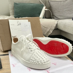 Christian Louboutin vita sneakers med nitar - Säljer ett par vita sneakers från Christian Louboutin i skinn med ikoniska röda sulor och coola nitar på tån. Skorna har snörning och en klassisk låg siluett. Perfekta för dig som vill sticka ut med exklusiv stil och edgy detaljer. Kontakta mig före du köper, jag reser ganska myckey