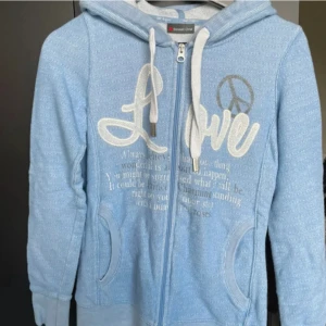 Ljusblå hoodie från Street One - Ljusblå hoodie med dragkedja från Street One. Tröjan har vita snören i huvan, broderad text och ett peace-märke framtill. Mjuk bomullskänsla och långa ärmar. Perfekt för en avslappnad stil.