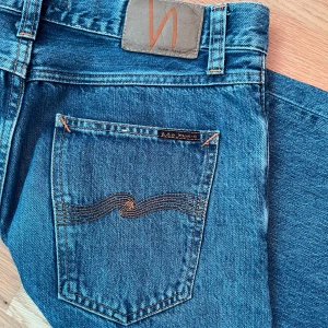 Blå jeans från Nudie Jeans - Säljer ett par klassiska blå jeans från Nudie Jeans med snygga kontrastsömmar och ikonisk brodyr på bakfickorna. Modellen har fem fickor, raka ben och normal passform. Perfekta för dig som gillar stilrena och tidlösa jeans. Storlek: Bredd 30, Längd 34. Original pris 1300 kr, andvända fem gånger.