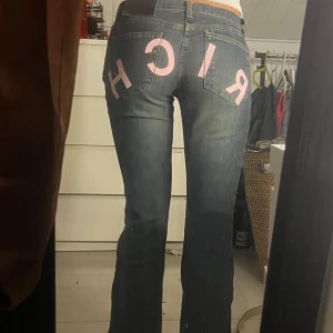 Blå bootcut jeans med rosa tryck - Unika blå jeans med bootcut passform och coolt rosa 'RICH'-tryck över bakfickorna. Jeansen har klassisk femficksdesign, knappgylf och är tillverkade i denim. Perfekta för dig som vill sticka ut med en statement-detalj.