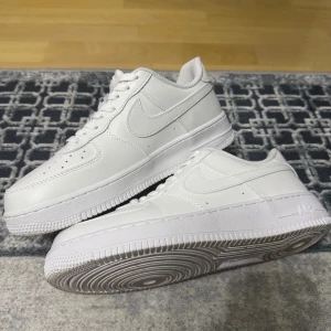 Nike Air Force 1  - Klassiska Nike Air Force 1 sneakers i helvitt skinn med vit sula och snörning. Ikonisk design med rund tå, perforerade detaljer på tån och broderad Nike-logga bak. Perfekta för dig som gillar en clean och tidlös look. Använda 1 gång 