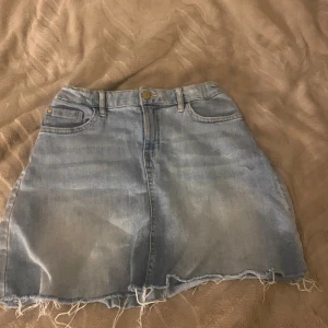 Ljusblå jeanskjol från Lindex - Säljer en ljusblå jeanskjol från Lindex med råa, fransiga kanter och klassiska fickor både fram och bak. Kjolen har bälteshällor och knappstängning framtill. Perfekt för dig som gillar en avslappnad och trendig stil.