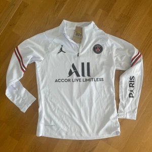 Vit PSG långärmad träningströja  - Snygg vit långärmad PSG fotbollströja från Jordan med dragkedja vid halsen, svarta och röda detaljer på ärmarna och tryck på bröstet och ryggen. Tillverkad i lätt och ventilerande funktionsmaterial. Storlek S. fortfarande ny med etikett