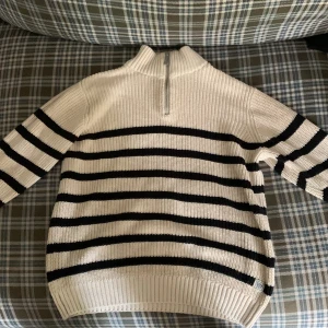 Randig stickad tröja från JINJUTS - Snygg stickad tröja från JINJUTS i storlek 134/140. Tröjan är vit med svarta horisontella ränder, har en hög krage och en halv dragkedja framtill. Perfekt för kyligare dagar och ger en klassisk look med modern touch.