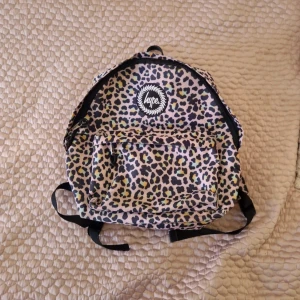 Leopardmönstrad ryggsäck från Hype - Cool ryggsäck från Hype med leopardmönster i svart, beige, gult och blått. Väskan har ett stort huvudfack, en praktisk ytterficka med dragkedja och justerbara axelremmar. Perfekt för skolan eller stan när du vill sticka ut lite extra.