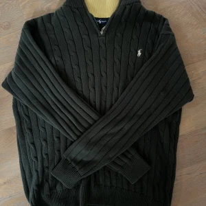 Kabelstickad Ralph Lauren tröja  - Mörkgrön kabelstickad tröja från Ralph Lauren med halv dragkedja och broderad logga på bröstet. Tröjan har ribbade muddar och en gul detalj på insidan av kragen. Perfekt inför vintern. Skriv om ni har funderingar !