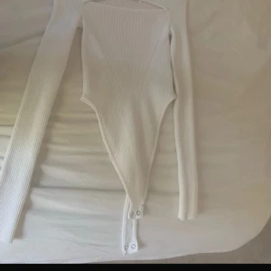 Vit ribbad långärmad bodysuit - Säljer en vit ribbad bodysuit med långa ärmar och knäppning nedtill. Toppen har en tight passform och är tillverkad i ett stretchigt material som sitter snyggt på kroppen. Perfekt att styla med jeans eller kjol för en clean look.