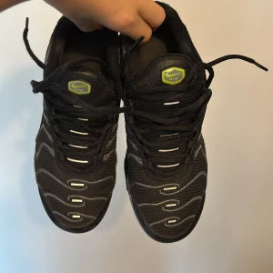 Nike Air Max Plus Tn i svart och neon - Nike Air Max Plus Tn sneakers i svart med detaljer i neon och gult. Skorna har klassisk Tn-logo bak, synliga air-bubblor i sulan och vågformade mönster på ovandelen. Ovandel i syntet och mesh, snörning framtill och robust yttersula med gula inslag.