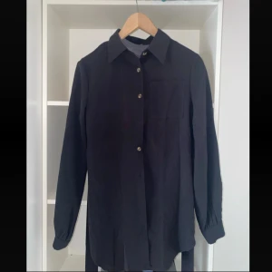 Svart skjorta med bälte och knappar - Stilren svart skjorta med klassisk krage, knäppning framtill och ett bröstficka. Skjortan har långa ärmar med manschett och ett bälte i midjan för extra detalj. Perfekt att slänga över en t-shirt för en chill look.