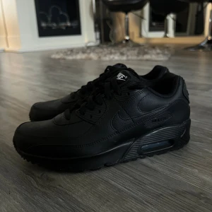 Nike Air Max 90 helsvarta sneakers - Nike Air Max 90 sneakers i helsvart skinn med klassisk chunky sula och synlig Air-enhet. Snygg och tidlös modell med snörning och diskreta detaljer. Perfekta för dig som gillar en clean och stilren look. Skorna är aldrig använda. Strlk 39 men passar även 38:)