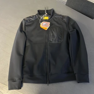Svart hybridjacka Parajumpers XL - Svart hybridjacka från Parajumpers i storlek XL. Jackan har quiltad rygg och axlar, bröstficka med dragkedja och hel dragkedja framtill. Materialmix med fleece och syntet ger en sportig och clean look. Perfekt för lager-på-lager under kyliga dagar. Den är precis nyköpt aldrig använd då den ej passade mig 