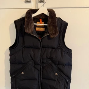 Svart dunväst från Parajumpers - Snygg svart dunväst från Parajumpers med mjuk brun fuskpäls på kragen och coola detaljer som snedställda fickor med knappar och dragkedja. Perfekt för lager-på-lager och har en sportig vibe med markerad krage och klassisk logga i nacken. Mycket sällsynt väst, slut producerad och slutsåld överallt. Priset kan diskuteras! 
