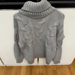 Grå polotröja med grov flätstickning - Säljer en chunky grå polotröja med breda flätstickade detaljer framtill och ribbad polokrage. Tröjan är stickad i ett tjockt garn och har långa ärmar med ribbade muddar. Perfekt för kalla dagar.