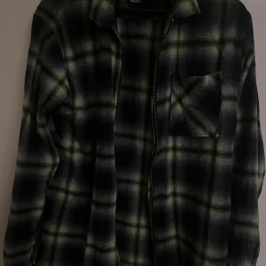 Rutig overshirt från FSBN i svart och grön - Säljer en oversized rutig overshirt från FSBN i svart och grön med dragkedja framtill och bröstficka. Skjortan har lång ärm och regular fit, perfekt för lager-på-lager-stil. Materialet känns mjukt och skönt mot huden.