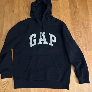 Svart GAP hoodie - Klassisk svart hoodie från GAP. Stor grå GAP-logga framtill, huva med snörning. Mjuk insida och ribbade muddar vid ärmslut och nederkant. 