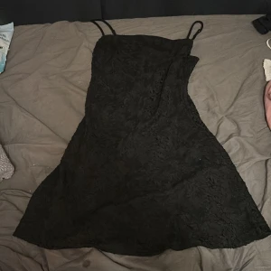 Svart kort klänning från BikBok - Snygg svart klänning från BikBok i kort modell med tunna axelband. Klänningen har ett diskret mönster i tyget och är tillverkad av 50% återvunnen polyester och 50% polyester. Perfekt för dig som gillar stilrena och trendiga plagg.