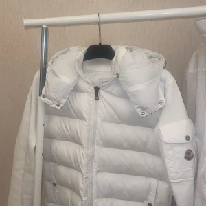 Vit Moncler  cardigan  - Snygg vit pufferjacka från Moncler med avtagbar huva och stickade ärmar. Jackan har dragkedja framtill, quiltad framsida och Moncler-logga på ärmen. Fickor med tryckknappar och coola detaljer gör den extra stilren. 