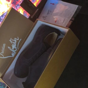 Mörkblå Christian Louboutin sneakers - Exklusiva mörkblå sneakers från Christian Louboutin med ikonisk röd sula. Skorna har snörning och är tillverkade i mocka med detaljer i skinn. Kommer med originalkartong och dustbag. Perfekta för dig som vill sticka ut med lyxig streetstyle.