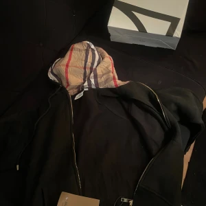 Svart hoodie med Burberry-mönstrad huva - Svart hoodie från Burberry med dragkedja och klassisk rutmönstrad huva i beige, rött och svart. Pris kan diskuteras i dm