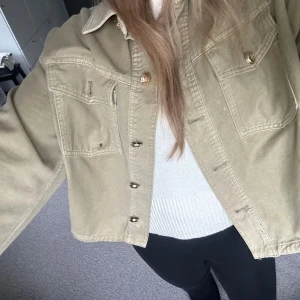 Grön jeansjacka med guldknappar - Säljer en beige jeansjacka med snygga guldknappar och två stora bröstfickor. Jackan har en loose passform och klassisk krage, perfekt att slänga över en hoodie eller t-shirt. Enkel och stilren design som funkar till det mesta.