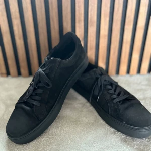 Svarta sneakers från Vagabond, strl 43 - Stilrena helsvarta sneakers från Vagabond i mocka med platt sula och rund tå. Skorna har klassisk snörning och en minimalistisk look som passar till det mesta. Perfekta för dig som gillar en clean och enkel stil. Lite tecken på slitage men annars i bra skick