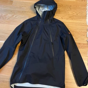  J.Lindeberg regnjacka - Snygg svart regnjacka från J.Lindeberg med huva och dragkedja snett över bröstet. Jackan har en minimalistisk look, diskret logga på ärmen och en praktisk ficka med dragkedja på ryggen. Perfekt för blöta dagar och enkel att matcha med allt. Storlek S men passar nog M oxå. använd 3 veckor Max. Orginalpris:3000:- 