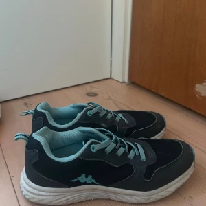 Svarta Kappa sneakers med turkosa detaljer - Säljer ett par svarta sneakers från Kappa med turkosa detaljer och snörning. Skorna har en vit sula och är tillverkade i en kombination av syntet och textil. Perfekta för dig som gillar sportig stil och vill ha bekväma skor till vardagen.