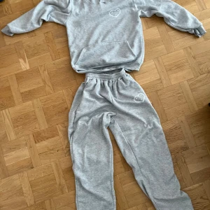 Carsicko tracksuit grå - Gråa tracksuit i bomull med elastisk midja och avslappnad passform. På vänster lår finns ett vitt tryck av ett hjärta med ansikte. Perfekta för chill dagar och matchar snyggt med hoodie i samma stil.